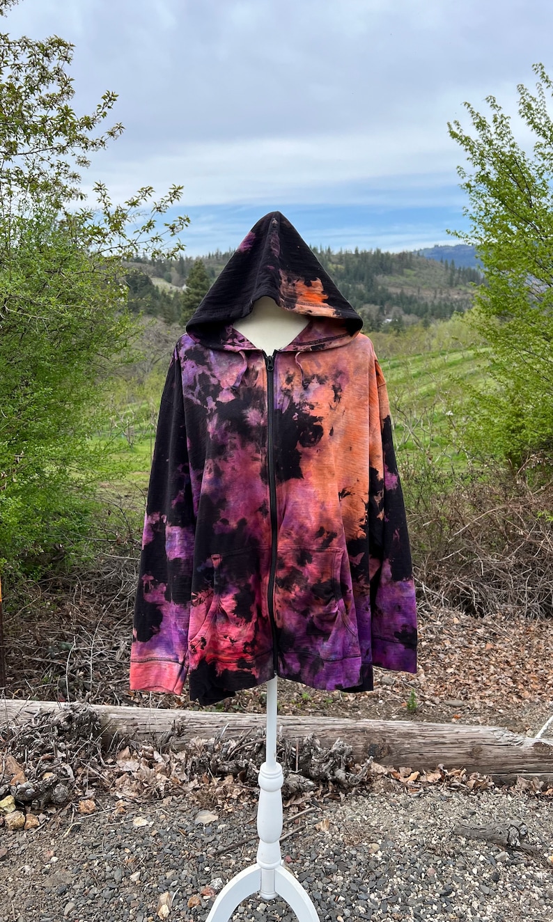 Op de afbeelding: Een zwarte hoodie met rits en een tie-dye patroon in tinten paars, oranje en zwart. De hoodie staat op een wit mannequin en heeft een capuchon.