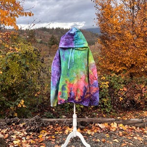 May include: A colourful tie-dye hoodie in shades of purple, blue, green, and pink is displayed on a white mannequin. The hoodie has a drawstring hood and a kangaroo pocket. The background features autumn foliage and a cloudy sky.