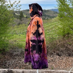 Op de afbeelding: Een tie-dye hoodie in zwart, oranje en paars met een capuchon met trekkoord. De hoodie staat op een wit mannequin tegen een achtergrond van groene bomen en een bewolkte lucht.