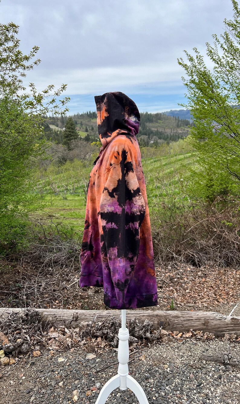 Op de afbeelding: Een tie-dye hoodie in zwart, oranje en paars met een capuchon met trekkoord. De hoodie staat op een wit mannequin tegen een achtergrond van groene bomen en een bewolkte lucht.