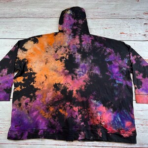 Op de afbeelding: Zwarte tie-dye hoodie met oranje, paarse en roze accenten. De hoodie heeft een capuchon met trekkoord en een ritssluiting.