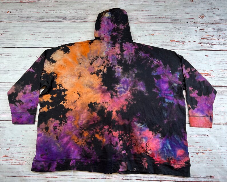 Op de afbeelding: Zwarte tie-dye hoodie met oranje, paarse en roze accenten. De hoodie heeft een capuchon met trekkoord en een ritssluiting.