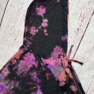 Op de afbeelding: Een zwarte tie-dye hoodie met een rits en een capuchon met trekkoord. De hoodie heeft een paars, roze en oranje tie-dye patroon.