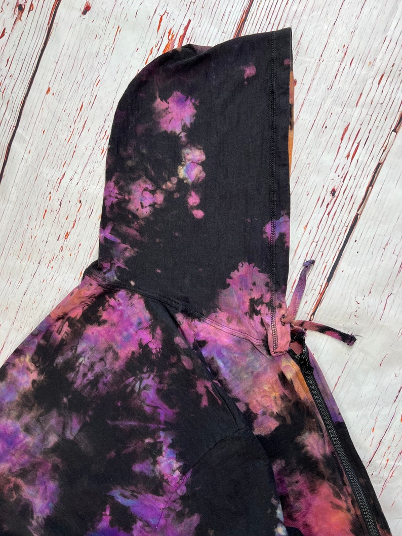 Op de afbeelding: Een zwarte tie-dye hoodie met een rits en een capuchon met trekkoord. De hoodie heeft een paars, roze en oranje tie-dye patroon.