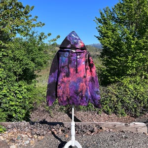 May include: A black tie-dye hoodie with purple, pink, and blue accents. The hoodie is on a white mannequin stand against a backdrop of green trees and a blue sky.