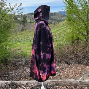 Op de afbeelding: Een zwarte hoodie met een tie-dye patroon in tinten roze, paars en blauw. De hoodie staat op een wit mannequin en is gefotografeerd buiten tegen een achtergrond van bomen en een bewolkte lucht.