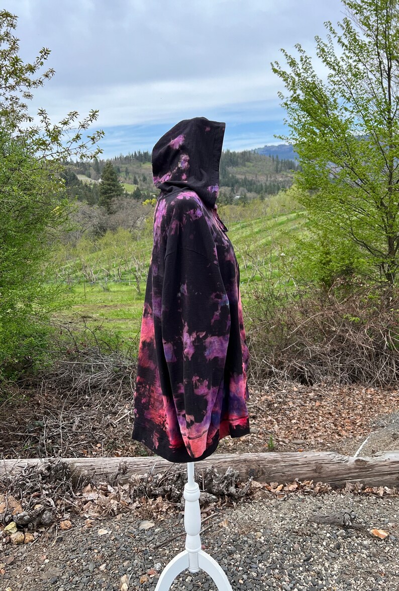 Op de afbeelding: Een zwarte hoodie met een tie-dye patroon in tinten roze, paars en blauw. De hoodie staat op een wit mannequin en is gefotografeerd buiten tegen een achtergrond van bomen en een bewolkte lucht.
