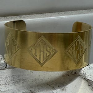 May include: A wide, gold-colored cuff bracelet. The bracelet features three diamond-shaped designs with etched initials. The cuff is open on the bottom and has a smooth, polished finish. The bracelet is a simple, yet elegant accessory.