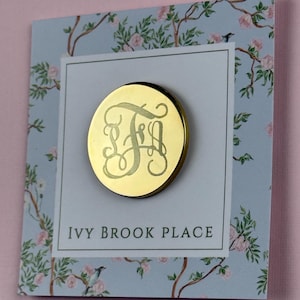 May include: A gold-colored round pin with an engraved monogram, displayed on a card with the text "IVY BROOK PLACE." The card has a floral design with pink flowers and green leaves on a light blue background.