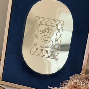 May include: A gold-toned framed mirror with a navy blue velvet backing. The mirror has an oval shape with a dragon design etched in the center. The frame has a gold border.