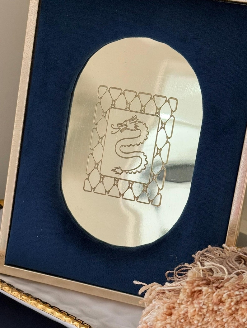 May include: A gold-toned framed mirror with a navy blue velvet backing. The mirror has an oval shape with a dragon design etched in the center. The frame has a gold border.