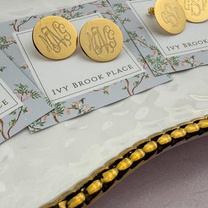 May include: Gold-toned circular stud earrings featuring an engraved monogram design. The earrings are displayed on rectangular cards printed with the text IVY BROOK PLACE and a floral pattern with birds, resting on a white tray with a gold scalloped edge.