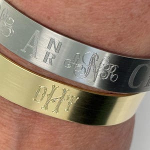 May include: Close-up of two engraved metal bracelets. The top bracelet is silver-toned with engraved initials and a script design. The bottom bracelet is gold-toned with engraved initials. Both bracelets are smooth and have a brushed metal finish.