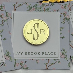 May include: A circular gold-toned metal pin featuring an engraved monogram with the letters J, S, and R. The pin is attached to a square white display card printed with the text IVY BROOK PLACE and a light blue floral pattern with pink flowers.