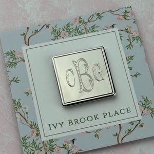 Peut inclure: Épingle à monogramme carrée argentée avec les lettres "CBG" dans une écriture élégante. L'épingle est posée sur une carte bleu clair avec le texte "IVY BROOK PLACE" et un motif floral avec des fleurs roses et des oiseaux.