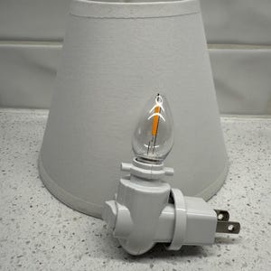 May include: A white night light with a clear, flame-shaped bulb and a white lampshade. The night light plugs directly into a wall outlet. The lampshade is a light gray color and has a tapered shape.