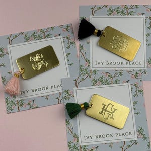 Puede incluir: Llaveros rectangulares dorados con iniciales grabadas y borlas de colores. Cada llavero está unido a una tarjeta con ilustraciones florales y de pájaros, y el texto "IVY BROOK PLACE".