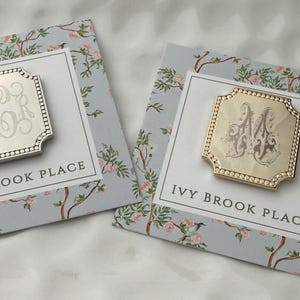 May include: Two square metal pins with beaded edges, one silver-toned with a script monogram and one gold-toned with a decorative letter M. Each pin is mounted on a light blue card featuring a floral pattern and the text IVY BROOK PLACE.