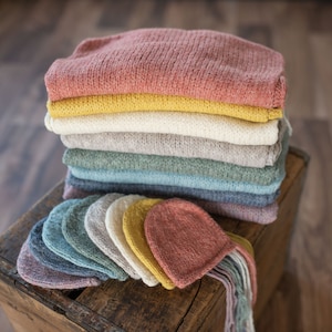 Op de afbeelding: Een stapel gebreide baby mutsen in verschillende kleuren, waaronder roze, geel, wit, groen, blauw en grijs. De mutsen liggen op een houten oppervlak.