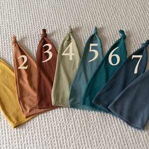 Puede incluir: Ocho gorros de bebé de tela de diferentes colores con los números del 1 al 8 impresos en ellos. Los gorros están dispuestos en una fila de izquierda a derecha.