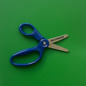 May include: A bright green folder with a pair of blue-handled scissors resting on top. The scissors have silver blades and the name "ANTHONY" printed on one blade. The folder is on a white, fluffy surface.