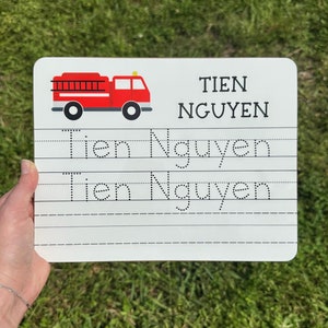 Puede incluir: Una pizarra blanca de borrado en seco con una ilustración de camión de bomberos rojo en la parte superior. La pizarra tiene líneas punteadas para trazar el nombre "Tien Nguyen".