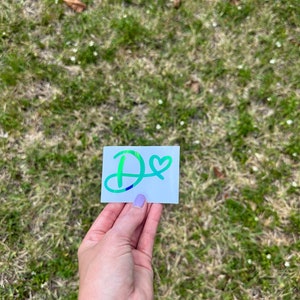 Può includere: Un adesivo bianco con una lettera "D" iridescente verde, blu e viola con un piccolo cuore accanto.