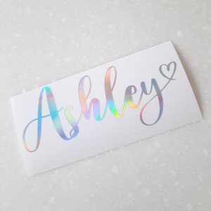 May include: A white sticker with the name "Ashley" written in a cursive font with a rainbow iridescent finish. A small heart is next to the name.