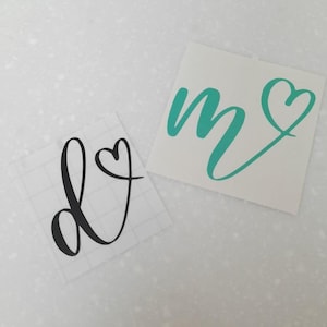 May include: Two vinyl decals, one with the letter "D" and a heart, and the other with the letter "M" and a heart. The "D" decal is black and the "M" decal is teal.