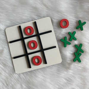 May include: A white wooden tic-tac-toe board with black lines and three red wooden 'O' pieces. There are four green wooden 'X' pieces off to the side.