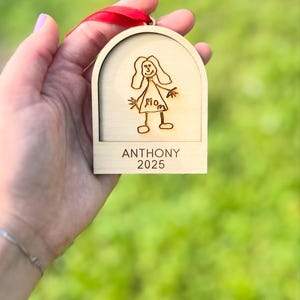 May include: A wooden ornament with a red ribbon for hanging. The ornament features a drawing of a person with the word "Mom" and the name "Anthony" with the year "2025" engraved below the drawing.