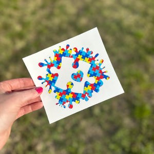 Puede incluir: Una calcoman&iacute;a de vinilo blanco con un dise&ntilde;o de pieza de rompecabezas azul, amarillo y rojo. La pieza del rompecabezas tiene un peque&ntilde;o coraz&oacute;n en el centro. La calcoman&iacute;a est&aacute; sobre un fondo blanco.
