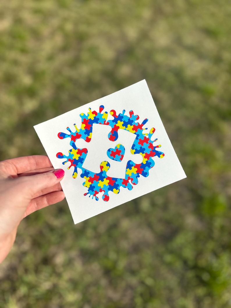 Puede incluir: Una calcoman&iacute;a de vinilo blanco con un dise&ntilde;o de pieza de rompecabezas azul, amarillo y rojo. La pieza del rompecabezas tiene un peque&ntilde;o coraz&oacute;n en el centro. La calcoman&iacute;a est&aacute; sobre un fondo blanco.