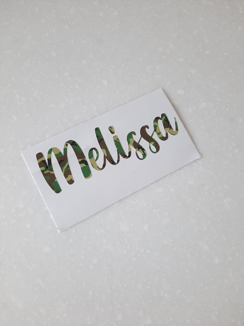 以下が含まれることがあります： 「Melissa」という名前が筆記体で書かれた白いステッカー。文字は切り抜かれており、緑、茶色、ベージュの迷彩柄で塗りつぶされています。