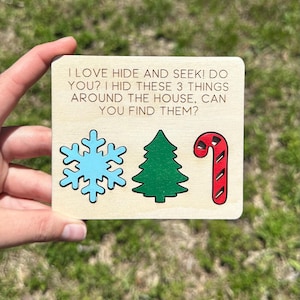 May include: Wooden game card with the text "I LOVE HIDE AND SEEK! DO YOU? I HID THESE 3 THINGS AROUND THE HOUSE. CAN YOU FIND THEM?" and three cutout shapes: a blue snowflake, a green Christmas tree, and a red and white candy cane.