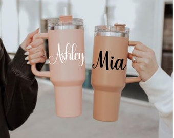 May include: Two personalized stainless steel travel mugs with handles. The mug on the left is pink and says "Ashley" in white script. The mug on the right is brown and says "Mia" in black script.