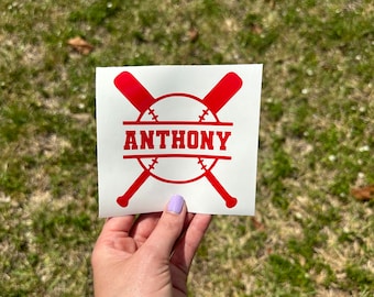 May include: A red baseball-themed sticker with the name "ANTHONY" in a bold, serif font. The design includes crossed baseball bats and a baseball shape. The sticker is held against a background of green grass.