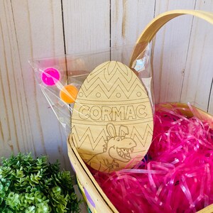 May include: A wooden Easter egg shaped craft with a dinosaur design and the name "CORMAC" written on it. The egg is in a basket with pink shredded paper and paint pots.