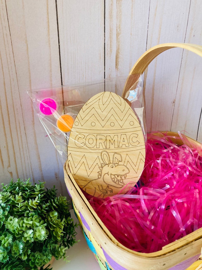 May include: A wooden Easter egg shaped craft with a dinosaur design and the name "CORMAC" written on it. The egg is in a basket with pink shredded paper and paint pots.
