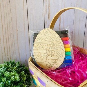 May include: A wooden Easter egg with a dinosaur design and the name "CORMAC" written on it. The egg is in a basket with pink and purple tissue paper and comes with a set of colored markers.