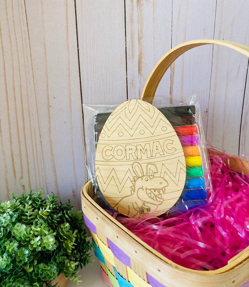 May include: A wooden Easter egg with a dinosaur design and the name "CORMAC" written on it. The egg is in a basket with pink and purple tissue paper and comes with a set of colored markers.