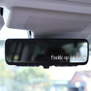 May include: A black rearview mirror with the text "Buckle up bitches" in white script, accompanied by a small heart. The mirror is mounted inside a vehicle, with the roof and windshield visible.