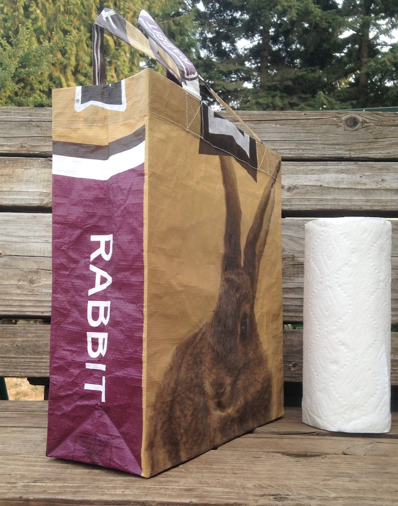May include: A large tote bag with a rabbit illustration. The bag is primarily tan with a large rabbit image and a vertical purple panel with the word "RABBIT" in white. The handles are black and white striped. A roll of white paper towels is next to the bag.