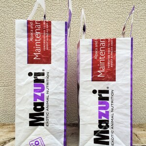 May include: Two white paper bags with handles, featuring the "Mazuri" logo in black and purple. The bags have a red and white label that reads "Alpaca and Maintenance". The bags are for exotic animal nutrition.