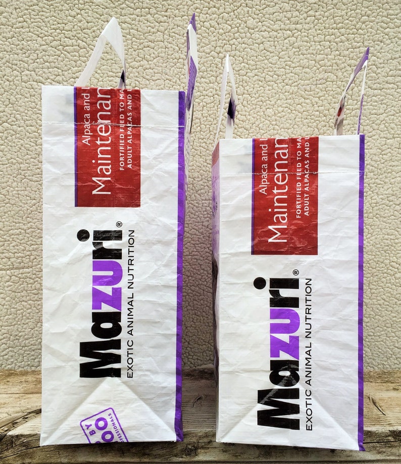 May include: Two white paper bags with handles, featuring the "Mazuri" logo in black and purple. The bags have a red and white label that reads "Alpaca and Maintenance". The bags are for exotic animal nutrition.