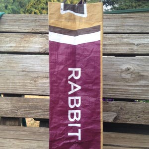 May include: A tall, rectangular bag with the word "RABBIT" in white capital letters. The bag has a burgundy lower section, a white stripe, and a tan upper section. The handles are striped.
