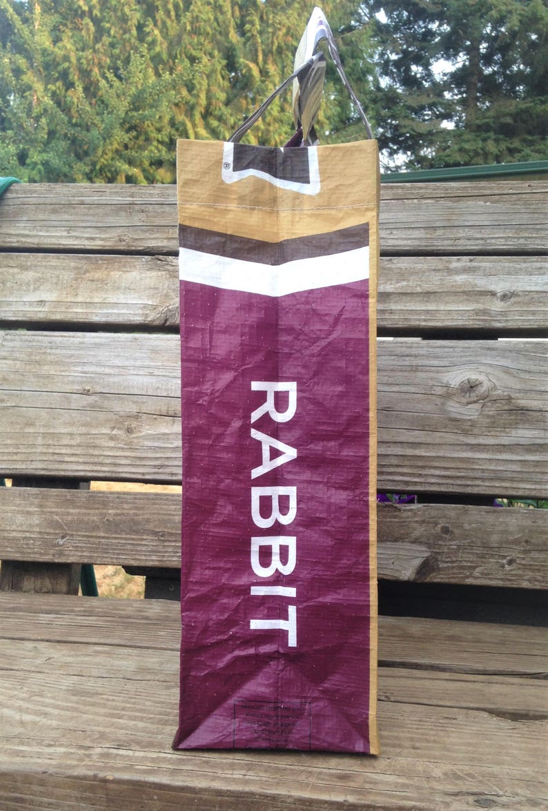 May include: A tall, rectangular bag with the word "RABBIT" in white capital letters. The bag has a burgundy lower section, a white stripe, and a tan upper section. The handles are striped.