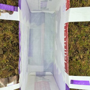 May include: An open, rectangular white bag with purple trim and handles, lying on the ground. The bag's interior is visible, and text is printed on the sides, including "Llamas" and "Certified Feed to Maintain Adult".