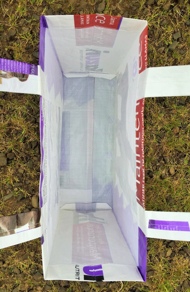May include: An open, rectangular white bag with purple trim and handles, lying on the ground. The bag's interior is visible, and text is printed on the sides, including "Llamas" and "Certified Feed to Maintain Adult".