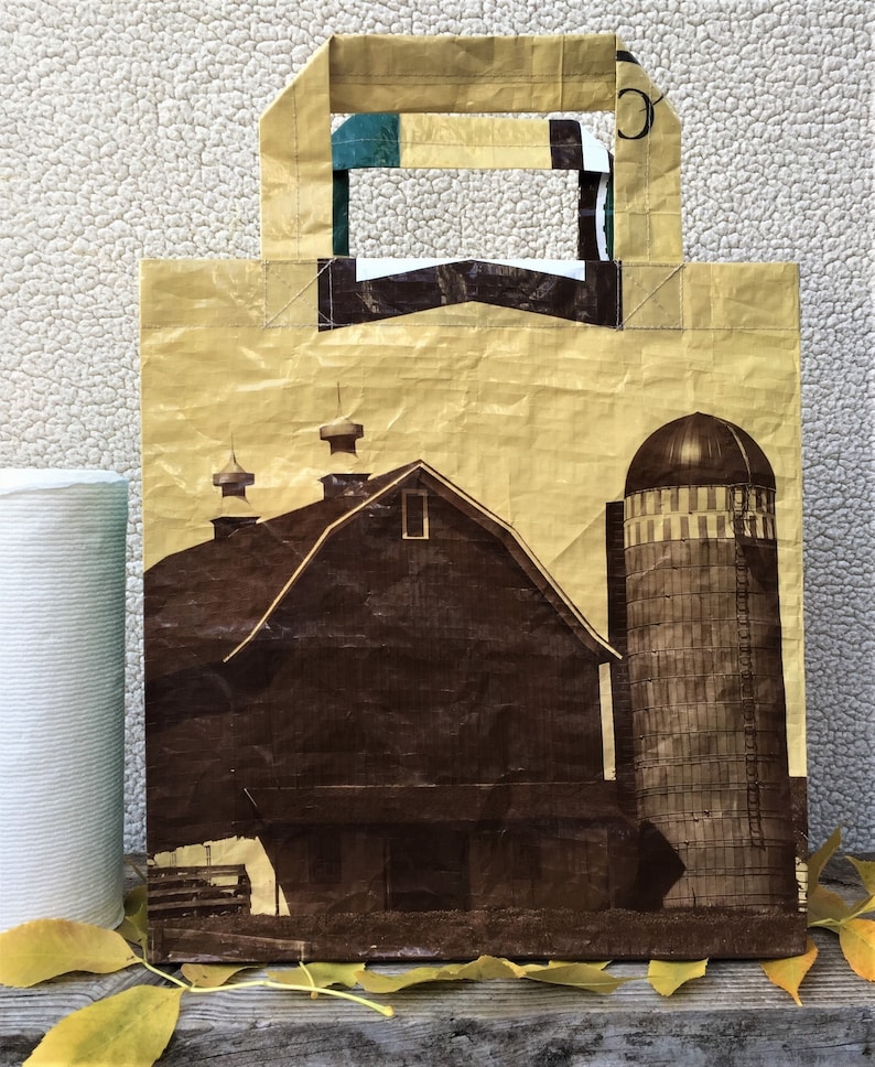 May include: A reusable shopping bag made from recycled materials. The bag features a brown and tan color scheme with a graphic of a brown barn and a silo. The bag has a brown handle.
